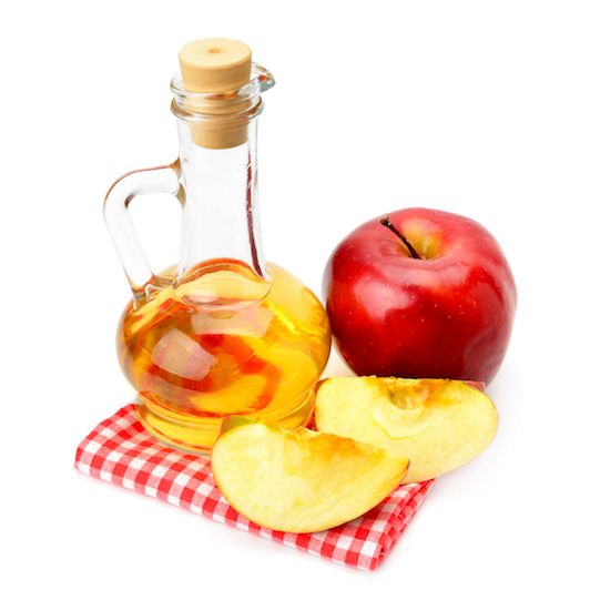 Äppelcidervinäger i liten flaska, rött äpple, två äppelklyftor, allt på en rödvit kökshandduk.