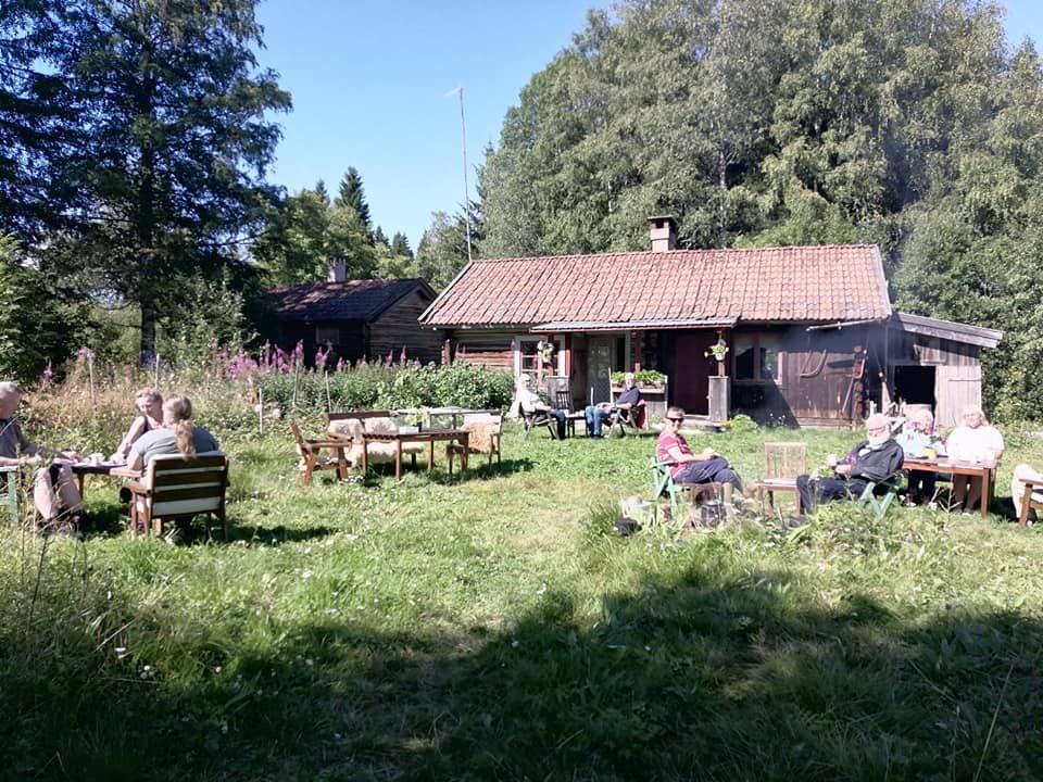 Personer sitter vid bord utanför en stuga i en solig trädgård.