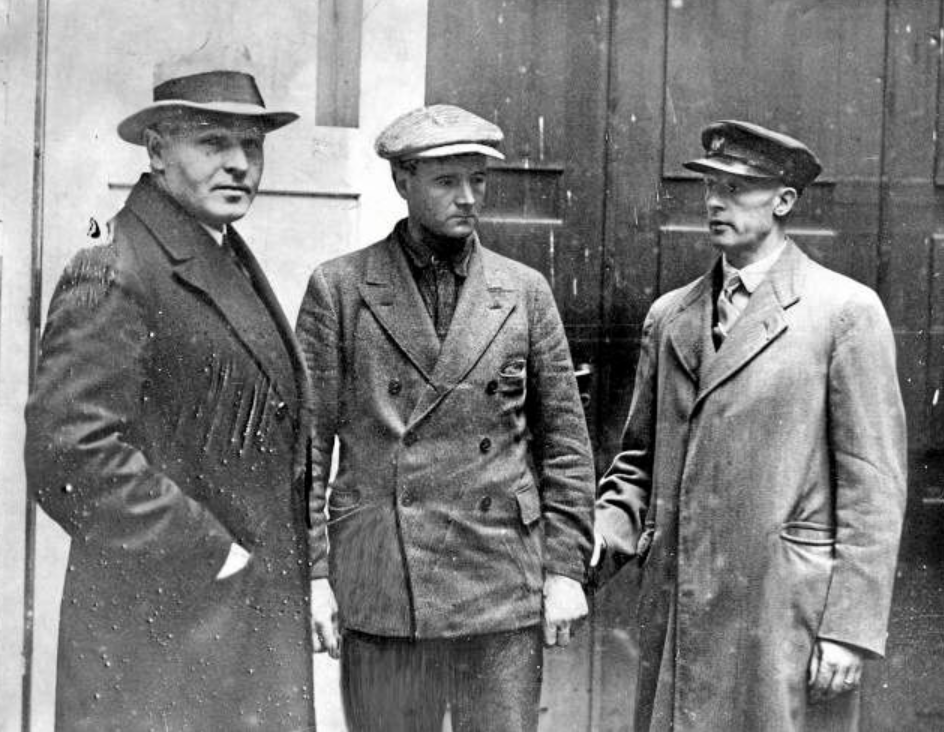 Gammalt svartvitt foto på tre män i rockar och hattar som står utomhus