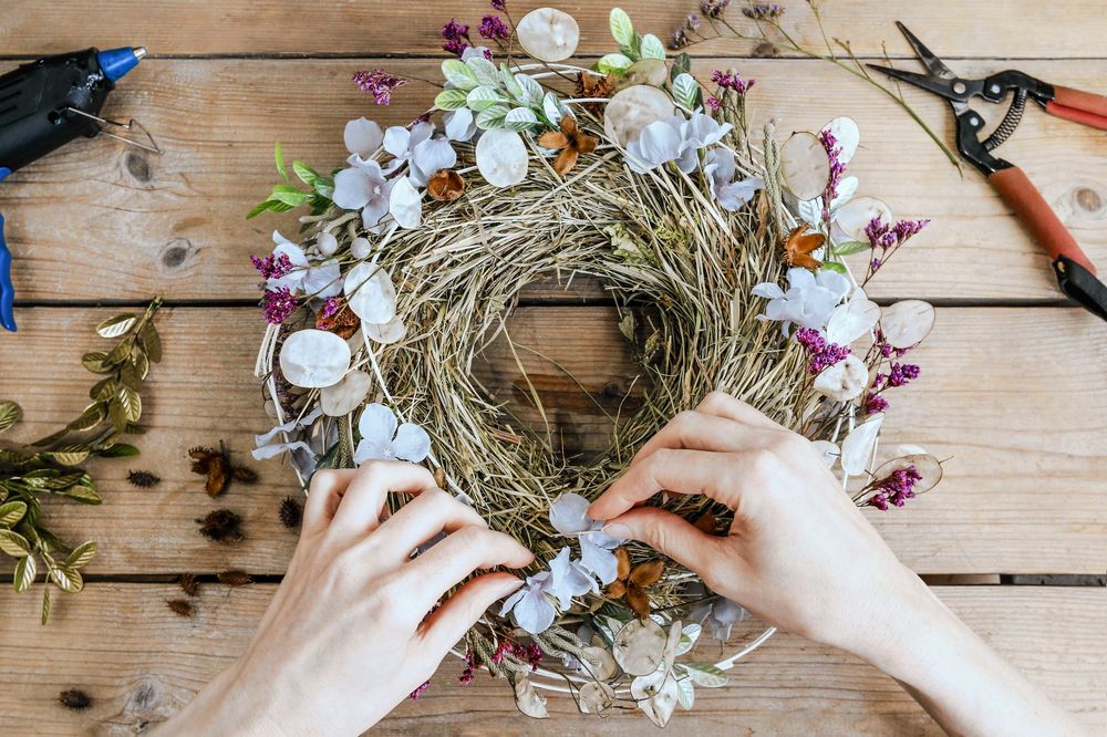 Händer skapar en blomkrans av torkade växter och blommor på ett träbord.