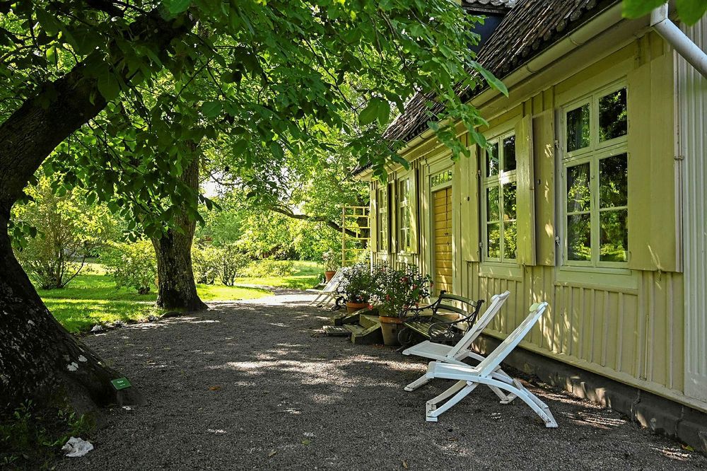 Grönskande träd och trädgård vid fasade av huset Hermina bodde i. Utanför huset står två vita trädgårdsstolar. 
