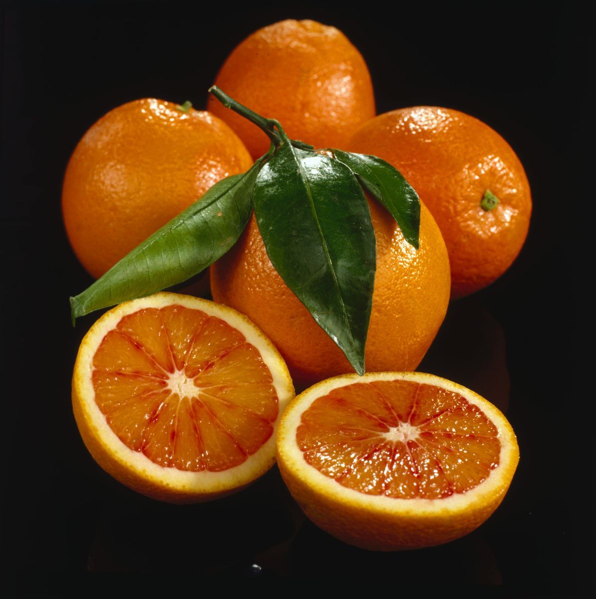 Blodapelsiner, hela och halverade, med gröna blad mot svart bakgrund