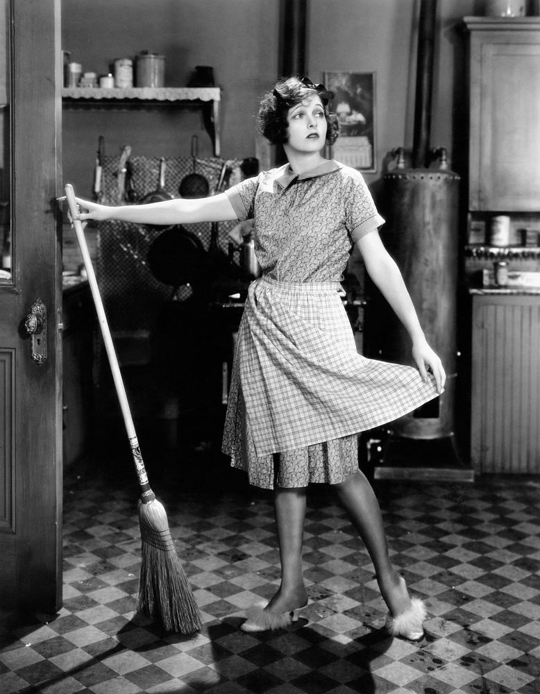 Svartvit bild på kvinna med sopkvast i gammeldags kök.