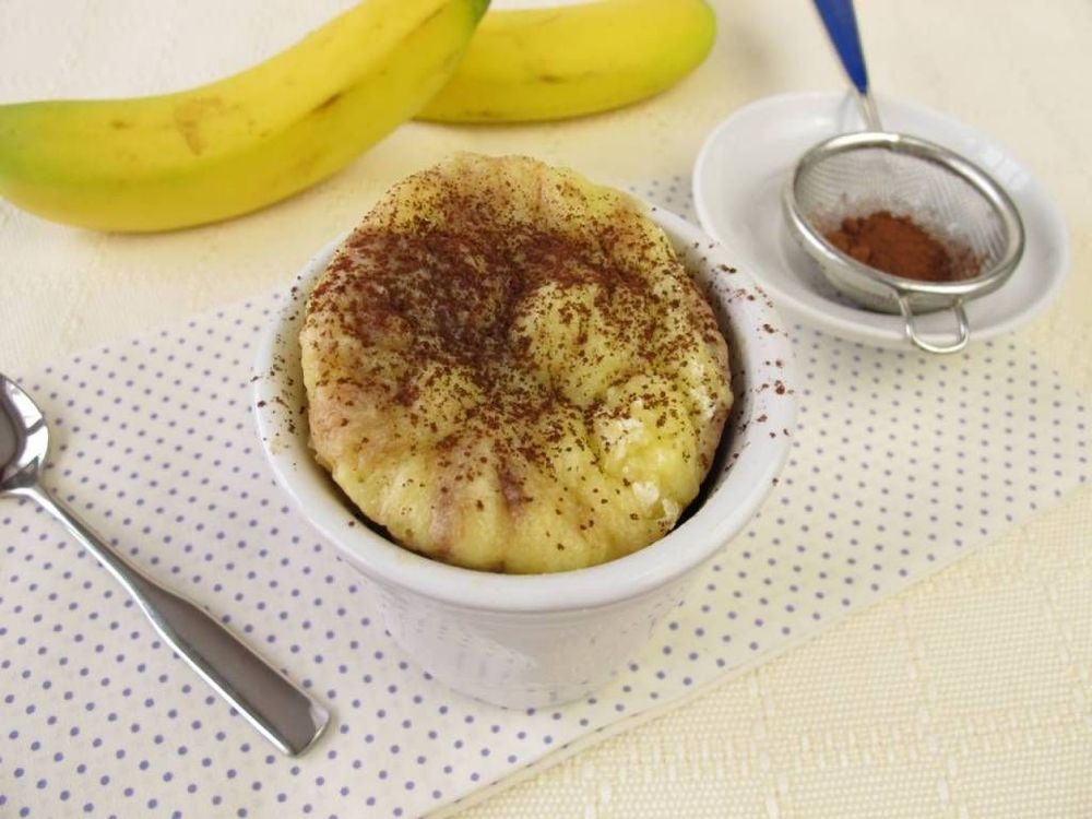 Banankaka i form med kanel strött på toppen. Vid sidan ligger två bananer och en skål med malen kanel i en tesil.