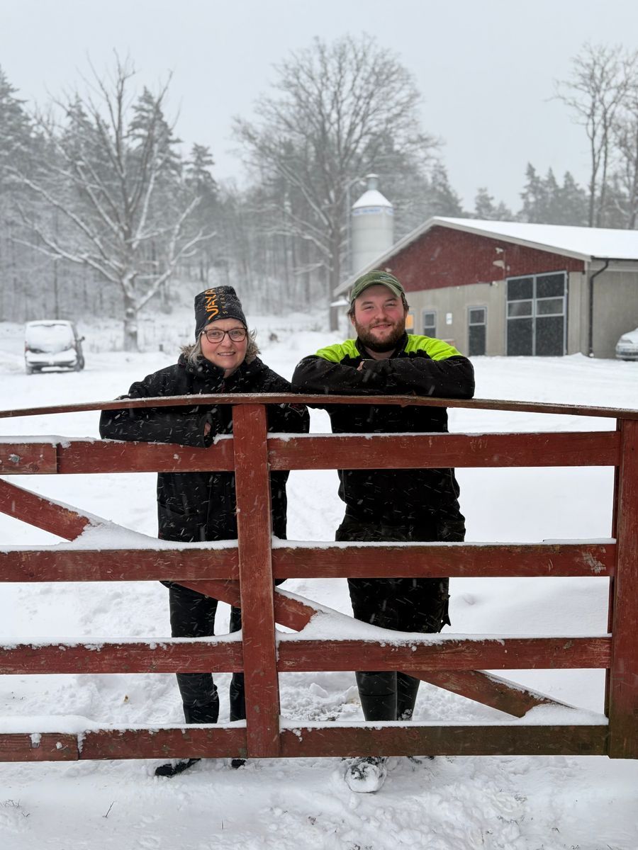 Klas Nilsson till höger och hans moster Anneli Karlsson till vänster vid en röd grind på en snöig gård med ladugård i bakgrunden