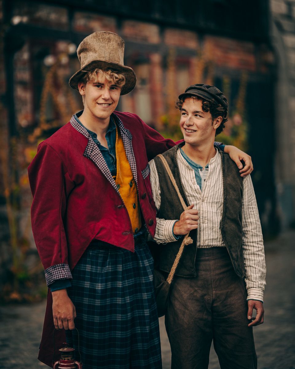 Två unga män i historiska kläder ler och står tillsammans på en stadsgata