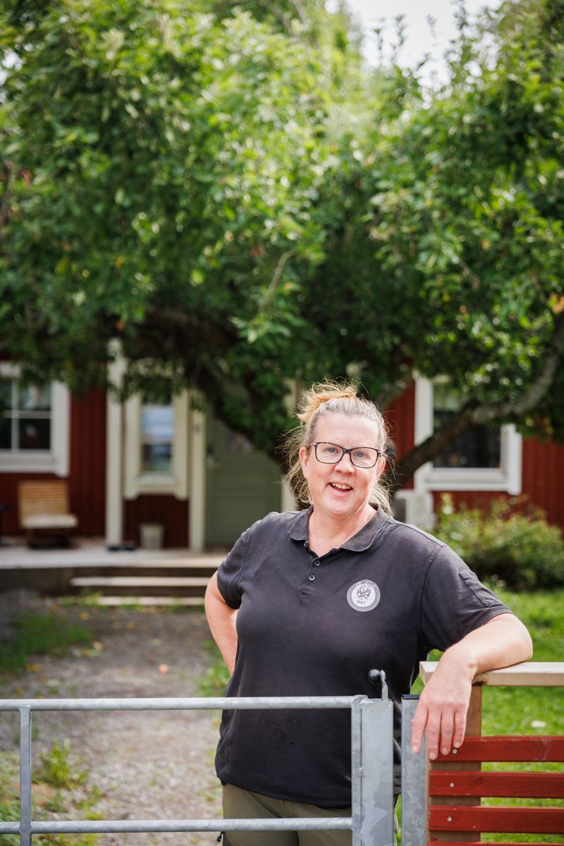 Leende kvinna vid grind framför rött hus och grönt trädgårdsträd