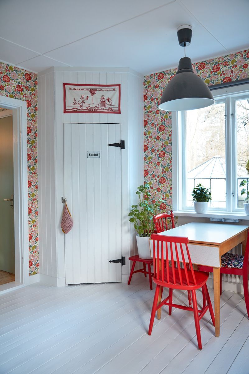 Ljust kökshörn med blommig tapet, vitt bord, röda stolar och vit trädörr