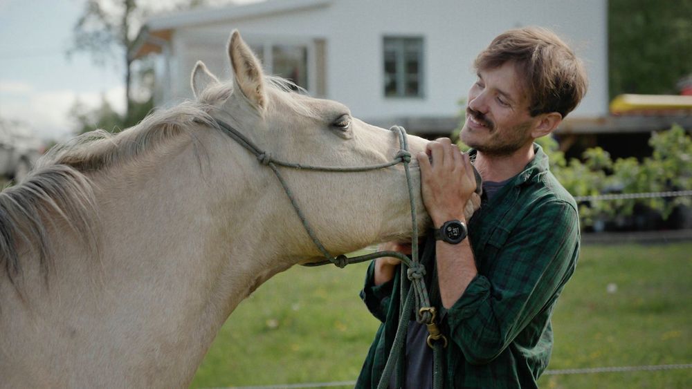Oskar Öhrström klappar hästen Ivory framför hus i somrig miljö.