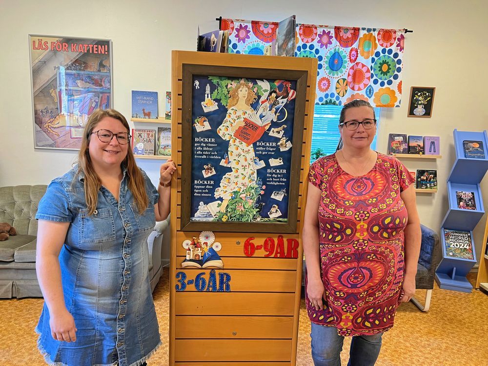 Två kvinnor står vid varsin sida av en bokhylla med barnböcker i ett bibliotek.