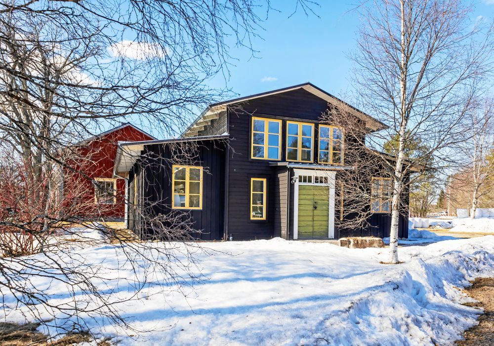 Svart träbyggnad med gula fönster, omgiven av snö och träd.