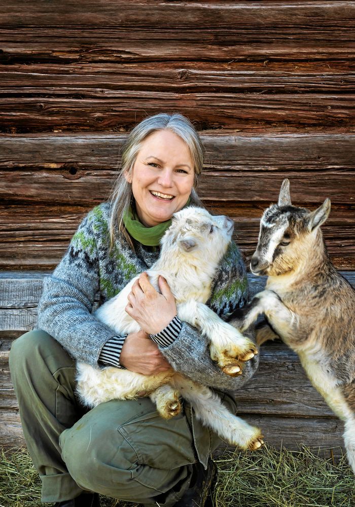 Emma sitter med två getter framför en trävägg, ler i grå och grön tröja.