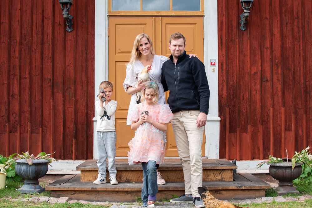 Familj på trappa framför rött trähus, barnen håller små djur