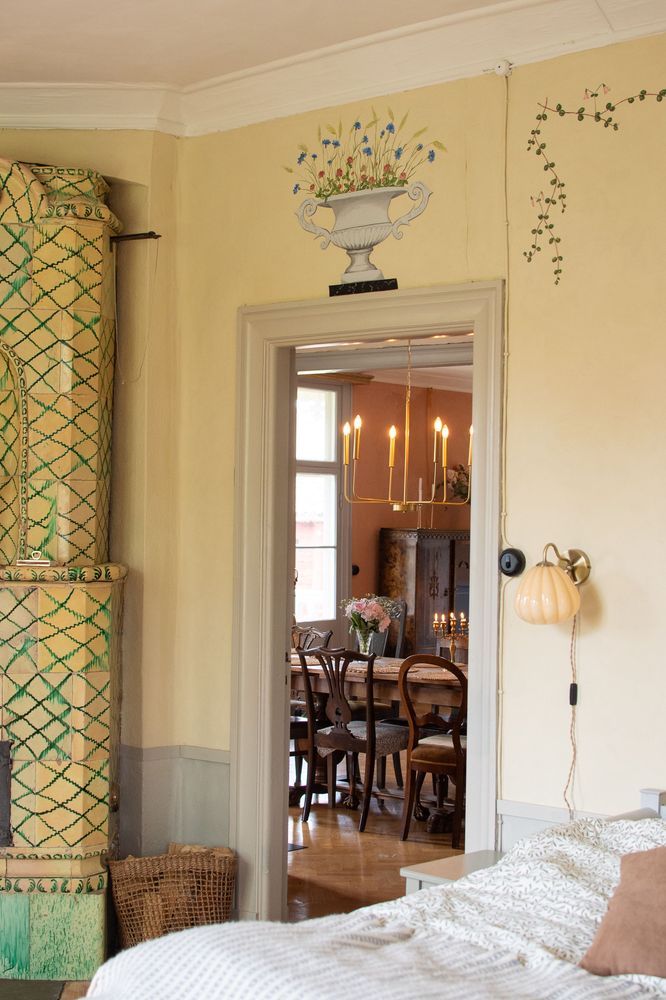 Sovrum med kakelugn och vy genom dörröppning mot matsal med tända ljus