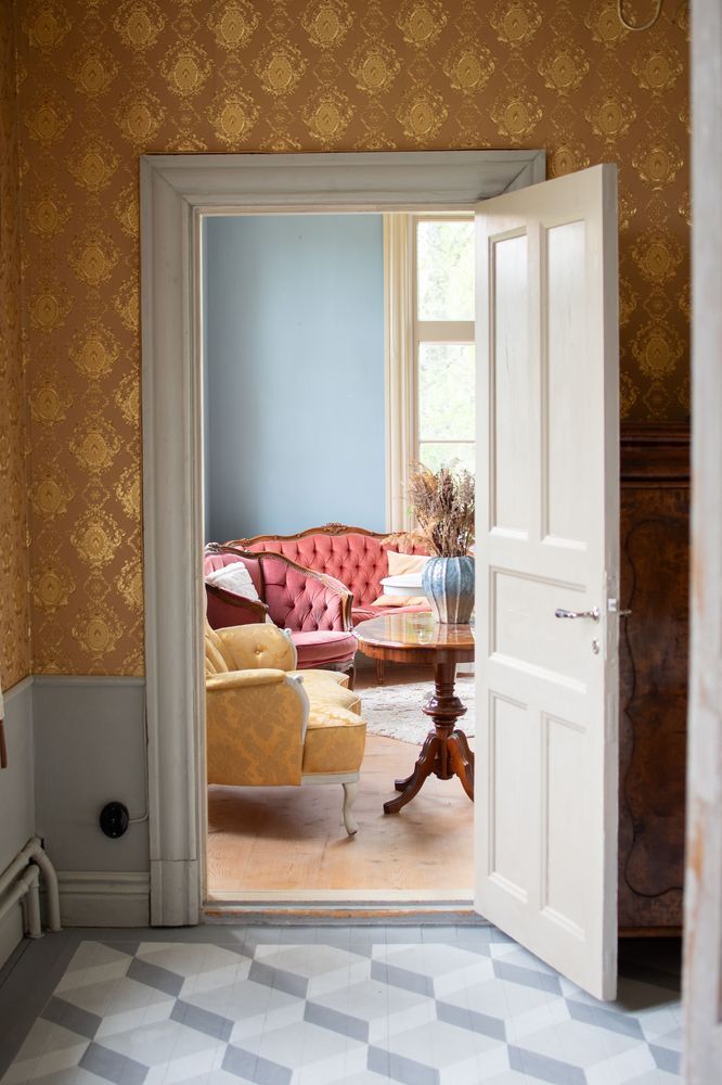 Öppen dörr mot vardagsrum med röd soffa, gult fåtölj och blommig tapet