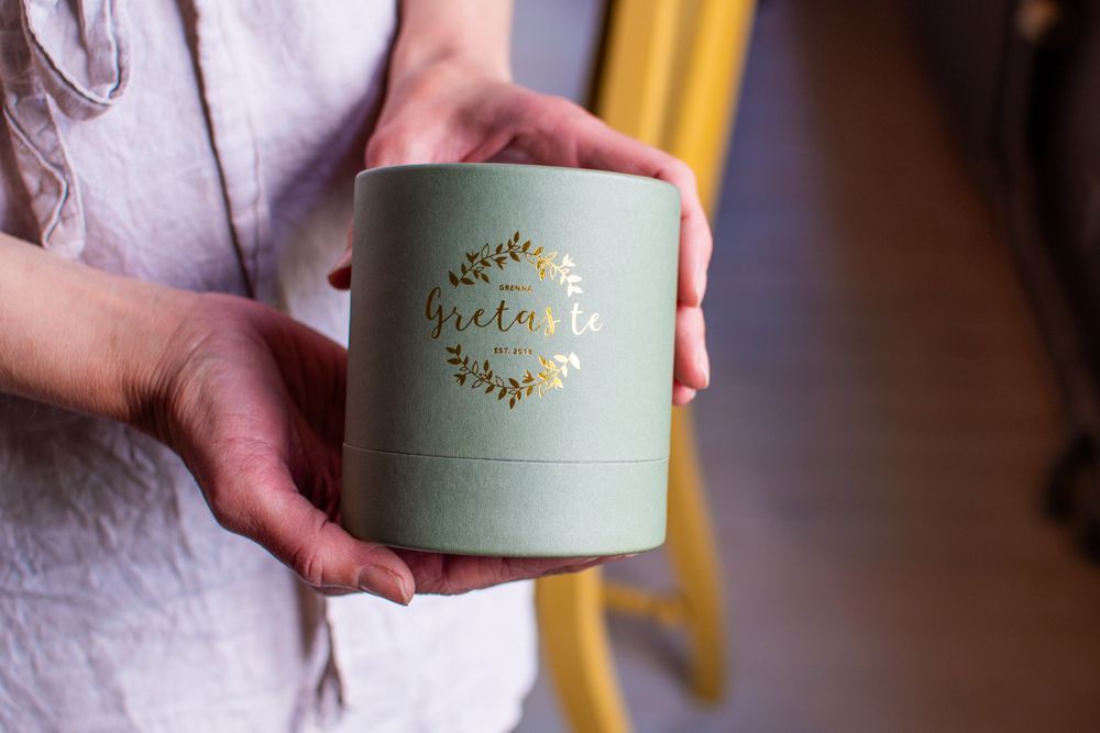 Kristine Yngfors håller en ljusgrön rund presentask med gyllene text och bladmotiv.