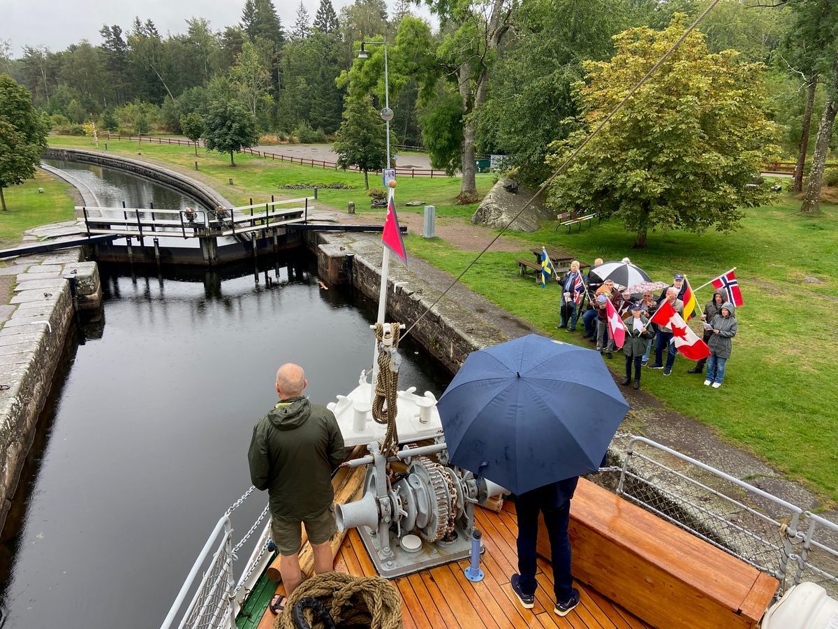 Båt i sluss i Göta kanal en regnig dag, människor på stranden vinkar med nordiska flaggor
