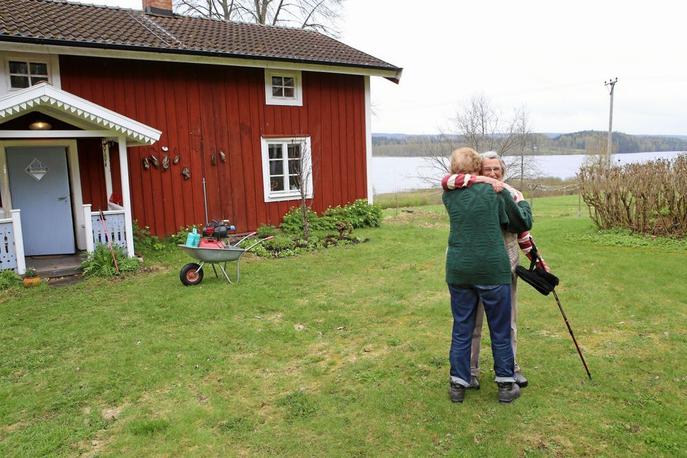 Två personer kramas framför ett rött hus med sjö i bakgrunden.