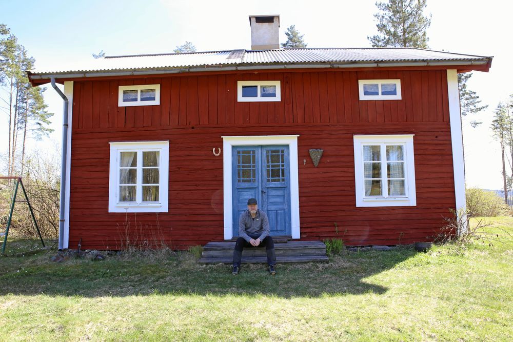 Man sitter på trappan till rött trähus i solsken.