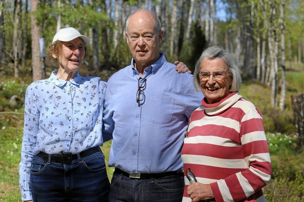 Tre äldre personer utomhus i skogen, omgivna av träd och soligt väder.