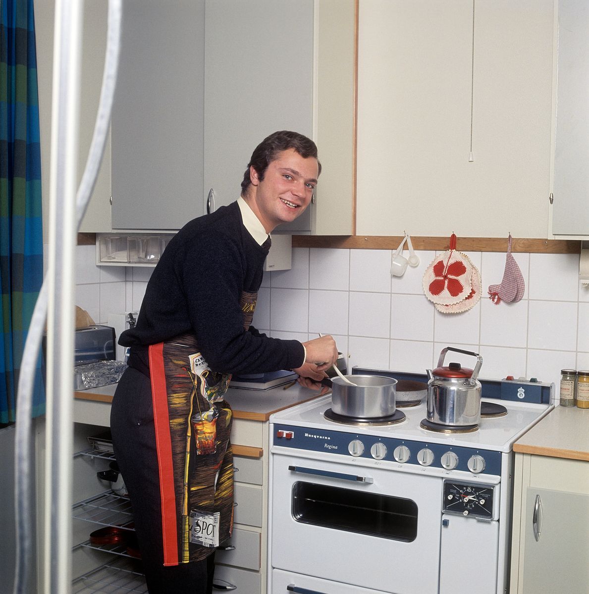 Leende kronprins Carl Gustaf lagar mat vid spis i ett kök med retroinredning