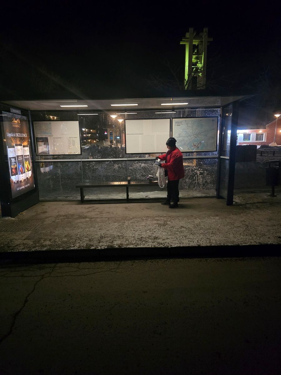 Person i röd jacka står vid busshållplats och läser tidtabell en mörk kväll