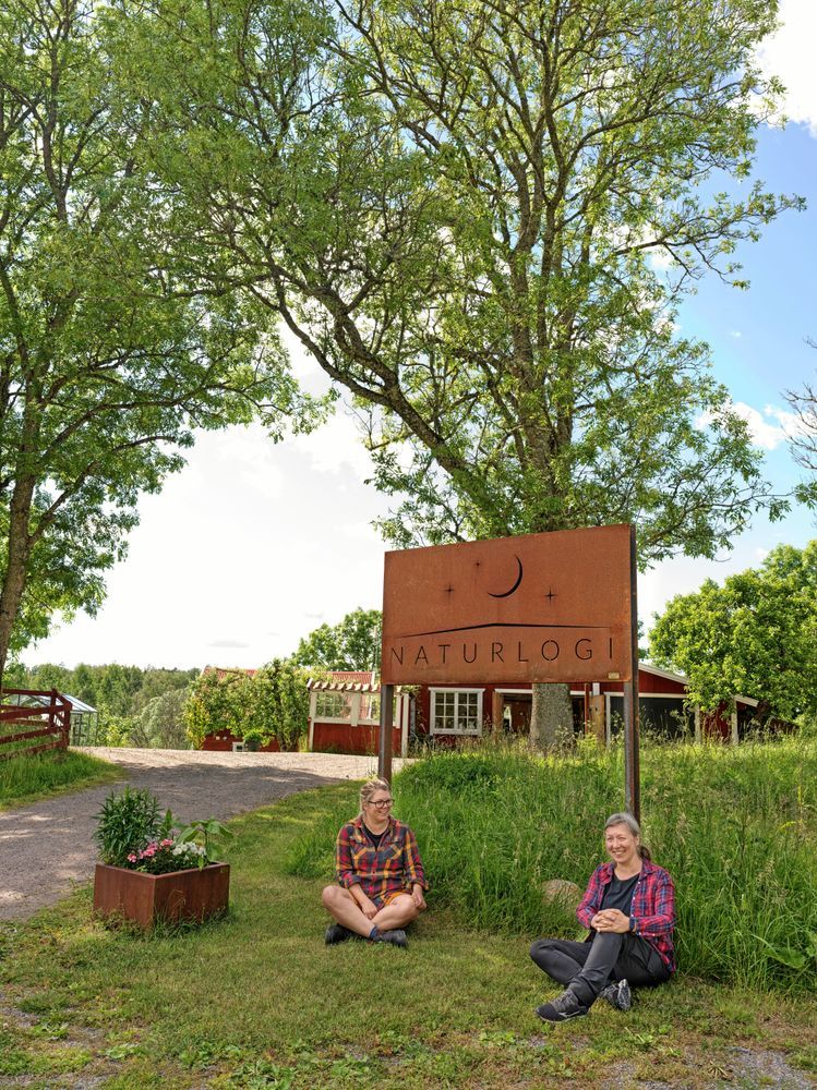 Linnéa Salbark och Josefine Espenkrona sitter framför en skylt med texten 'Naturlogi' vid ett naturskönt område.