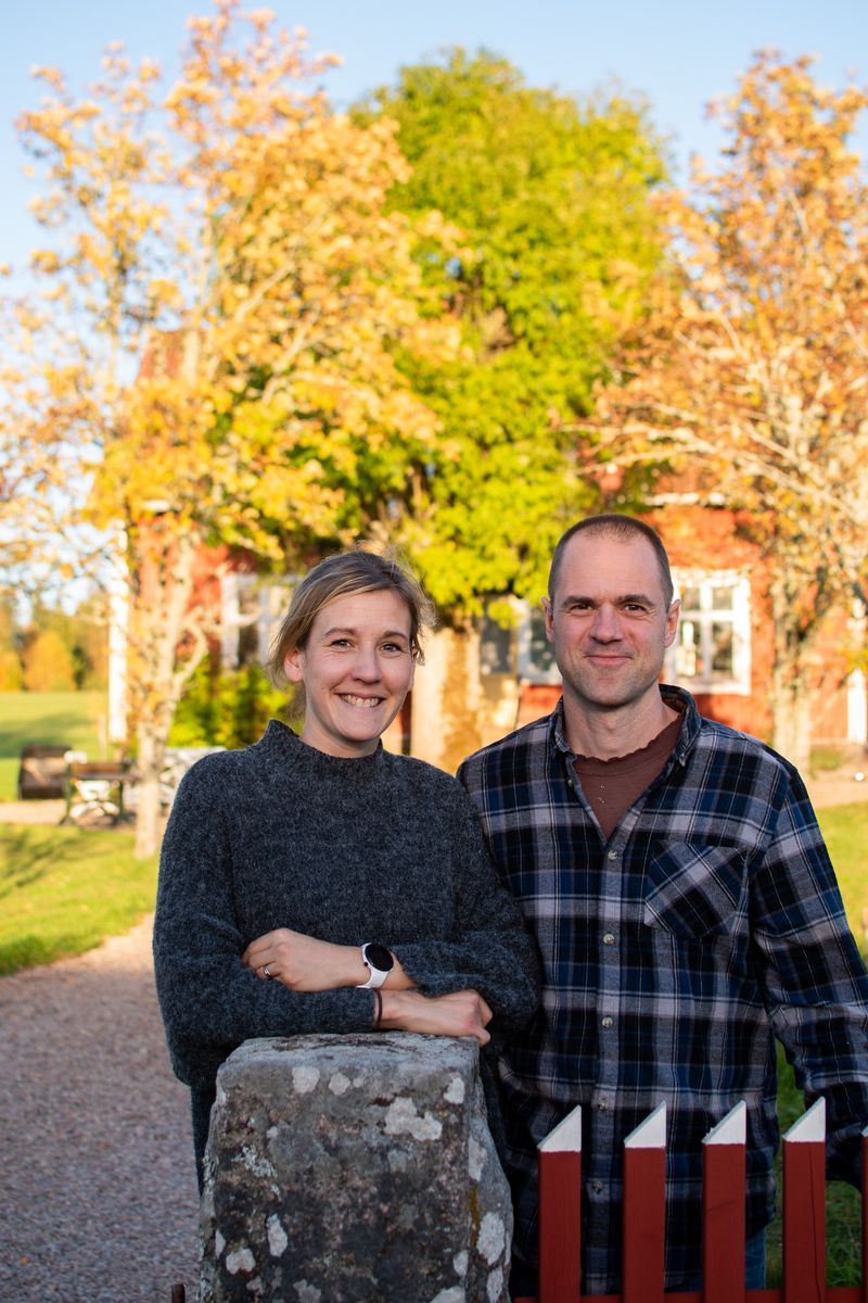 Josefine och Fredrik framför ett rött hus med höstträd i bakgrunden.