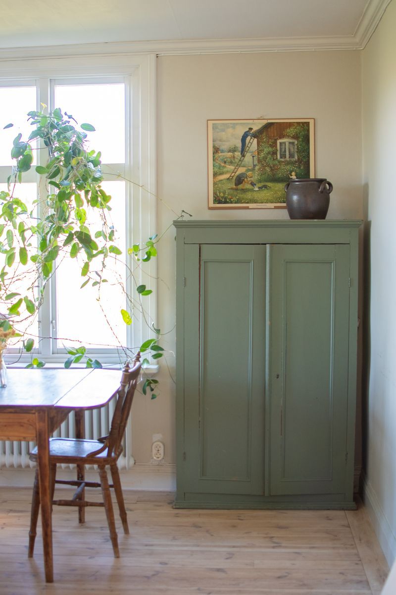 Grönt skåp och tavla i solbelyst rum med träbord och stolar.