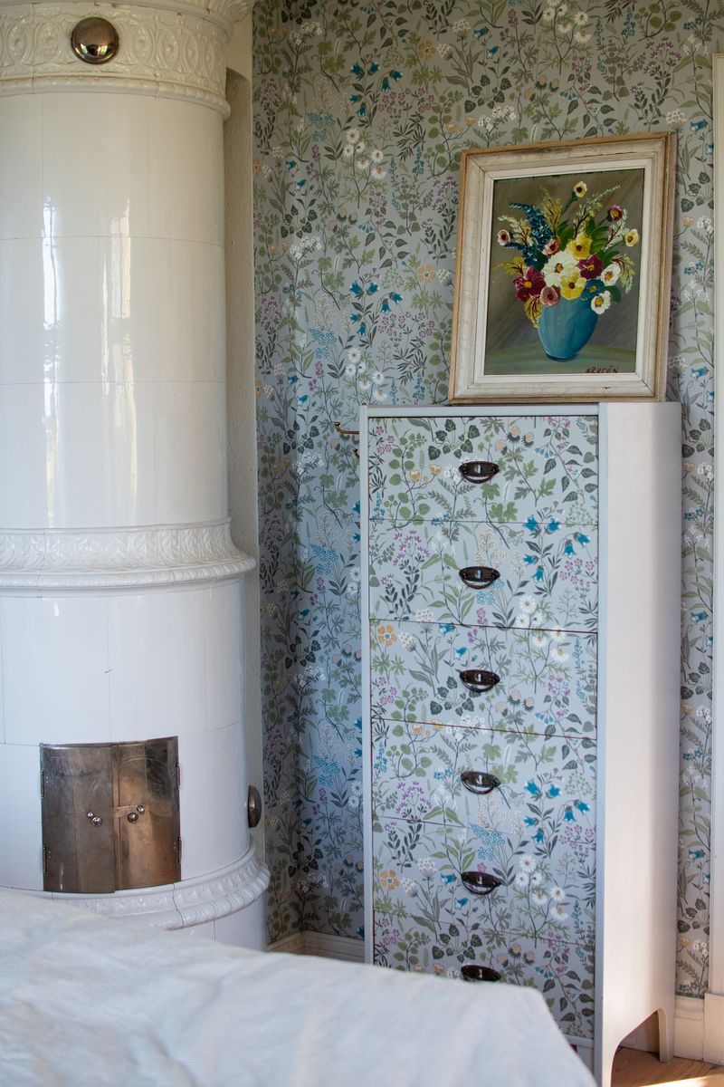 Kakelugn bredvid blommig byrå och tavla med blommor.