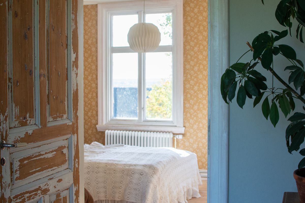 Sliten trädörr in till sovrum med beigeblommiga tapeter och säng med spetsöverkast.