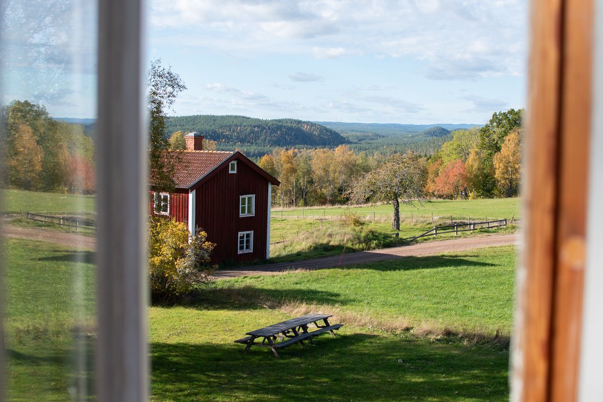 Utsikt från fönster mot röd stuga och höstlandskap.