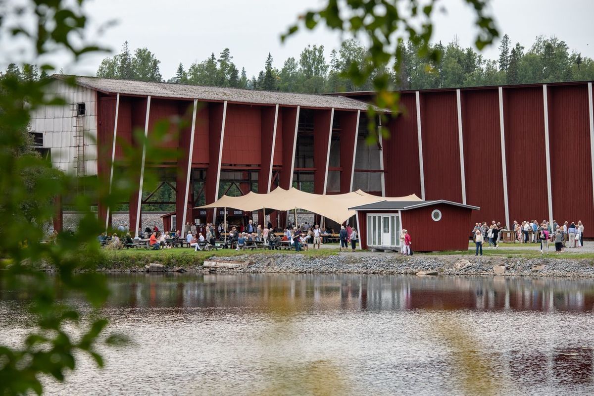 Människor samlade vid röd byggnad vid sjö, omgivna av skog