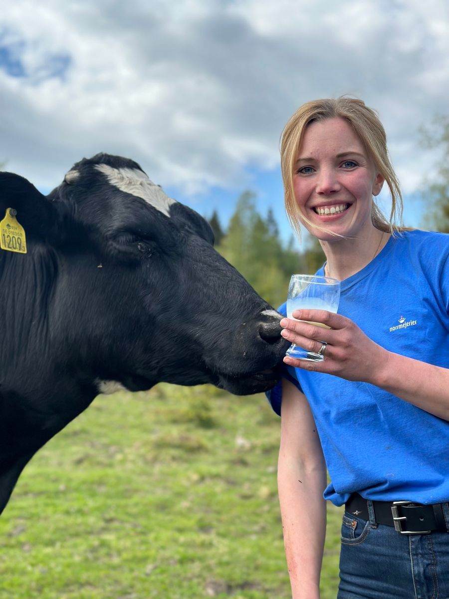 Leende kvinna i blå tröja håller ett glas mjölk bredvid en ko på grön äng