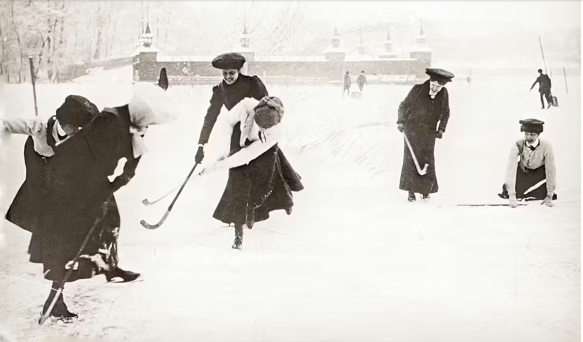 Svartvit historisk bild av kvinnor i långa kjolar som spelar hockey på isen