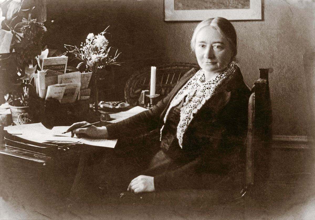 Äldre kvinna sitter vid skrivbord med papper och blommor i ett gammaldags rum