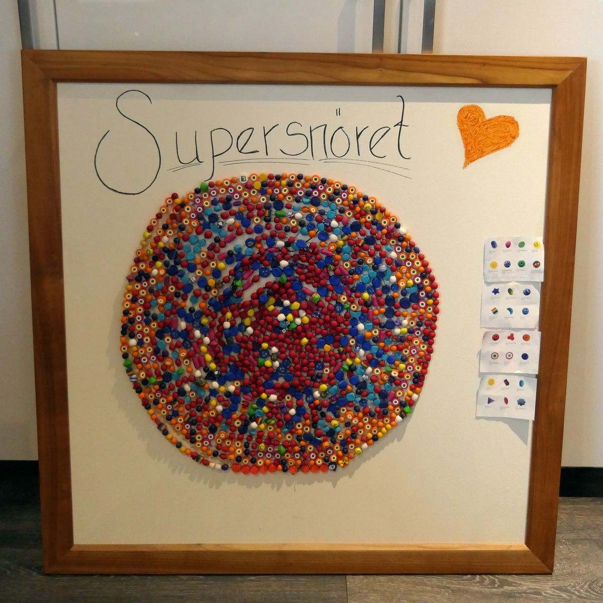 Tavla med texten ”Supersnöret” och en stor färgglad cirkel av pärlor