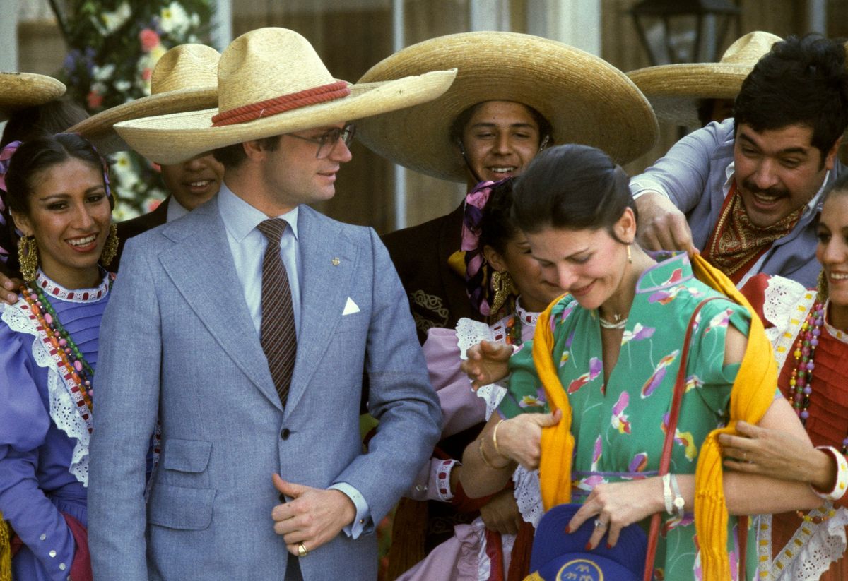 Kung Carl Gustaf i sombrero och blå kostym, drottning Silvia i ärmlös mönstrad klänning