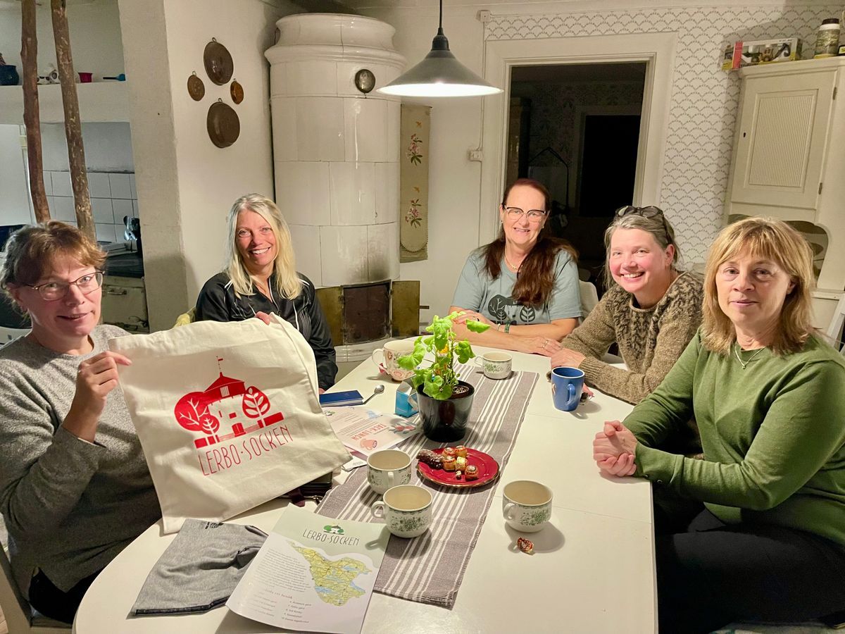 Fem leende kvinnor runt köksbord med fika och tygkasse med texten 'Lerbo socken'