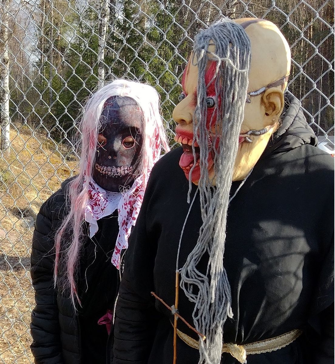 Två personer med. skrämmande masker framför staket i skogsmiljö.