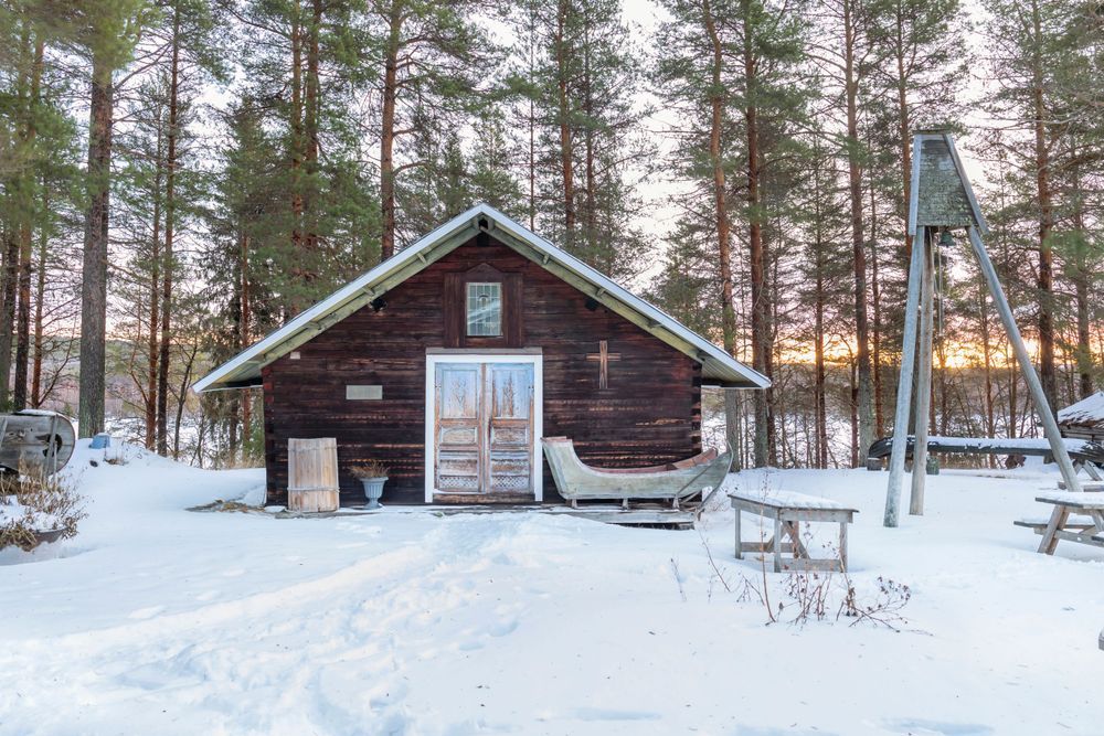 Trästuga omgiven av snö och tallar i vinterlandskap.