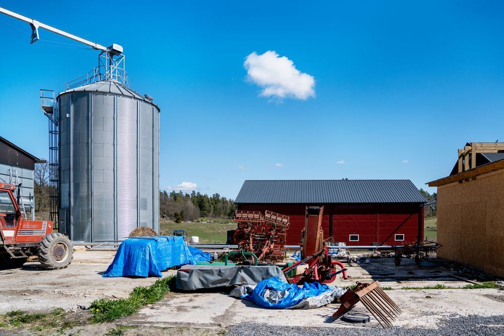Gård med metall­silo, röda lador och utspridda jordbruksmaskiner under blå himmel