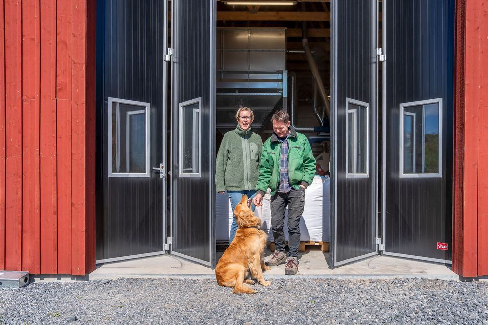 Thomas Björklund och Ida Lindberg Klausen med en hund står i dörröppningen till en röd lagerbyggnad