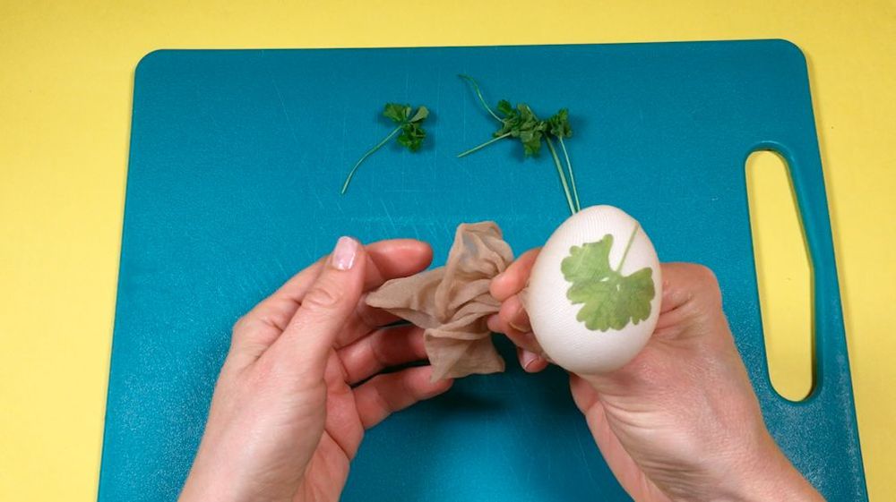 Händer dekorerar ett ägg med ett blad och nylonstrumpa på skärbräda