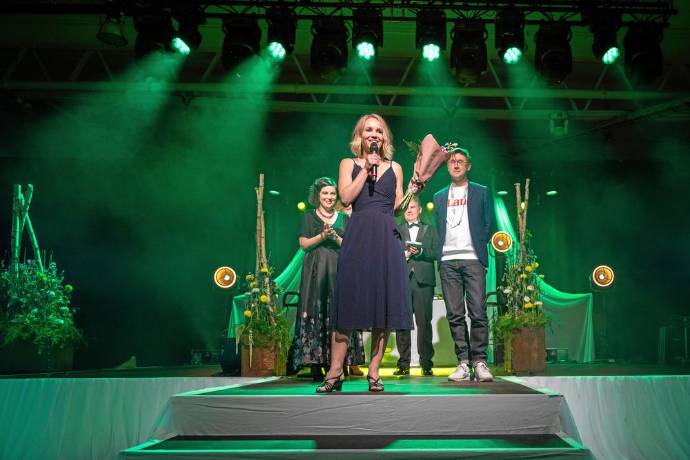 Kvinna på scen med mikrofon, tre personer i bakgrunden.