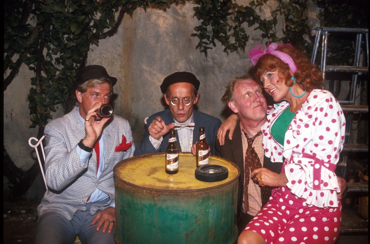 Grupp på fyra vid en grön tunna, dricker öl och pratar i en scenlik miljö.