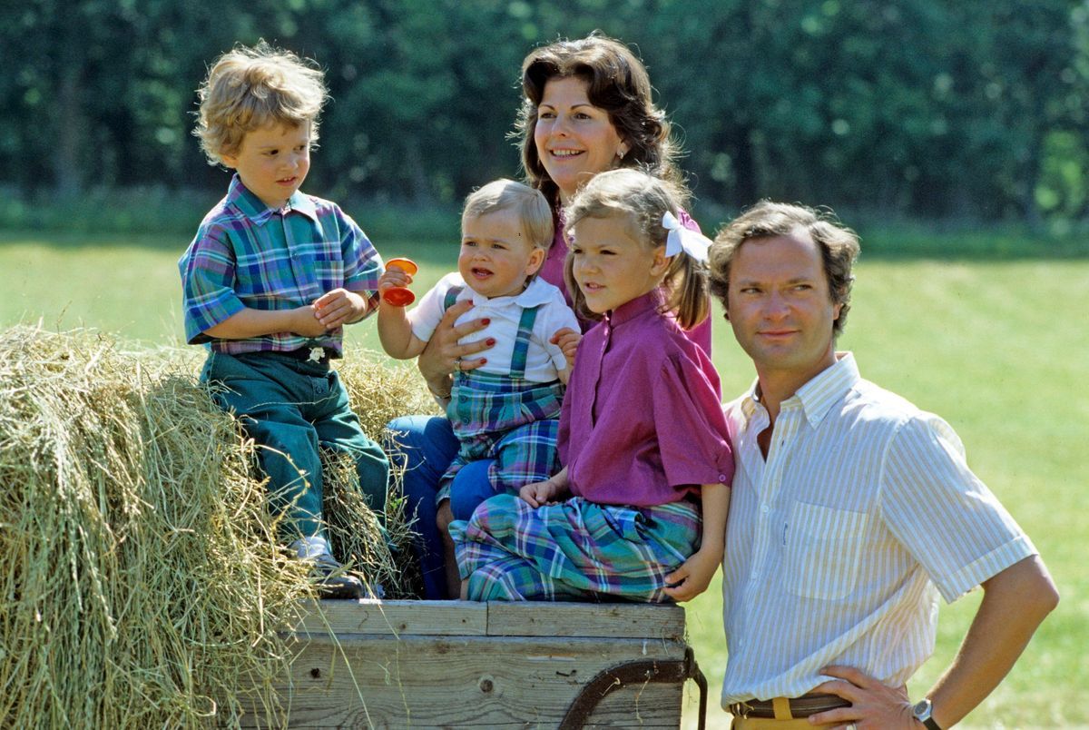 Kungafamiljen på Solliden Öland, drottningen och de tre småbarnen sitter på ett hölass, kungen står bredvid
1983