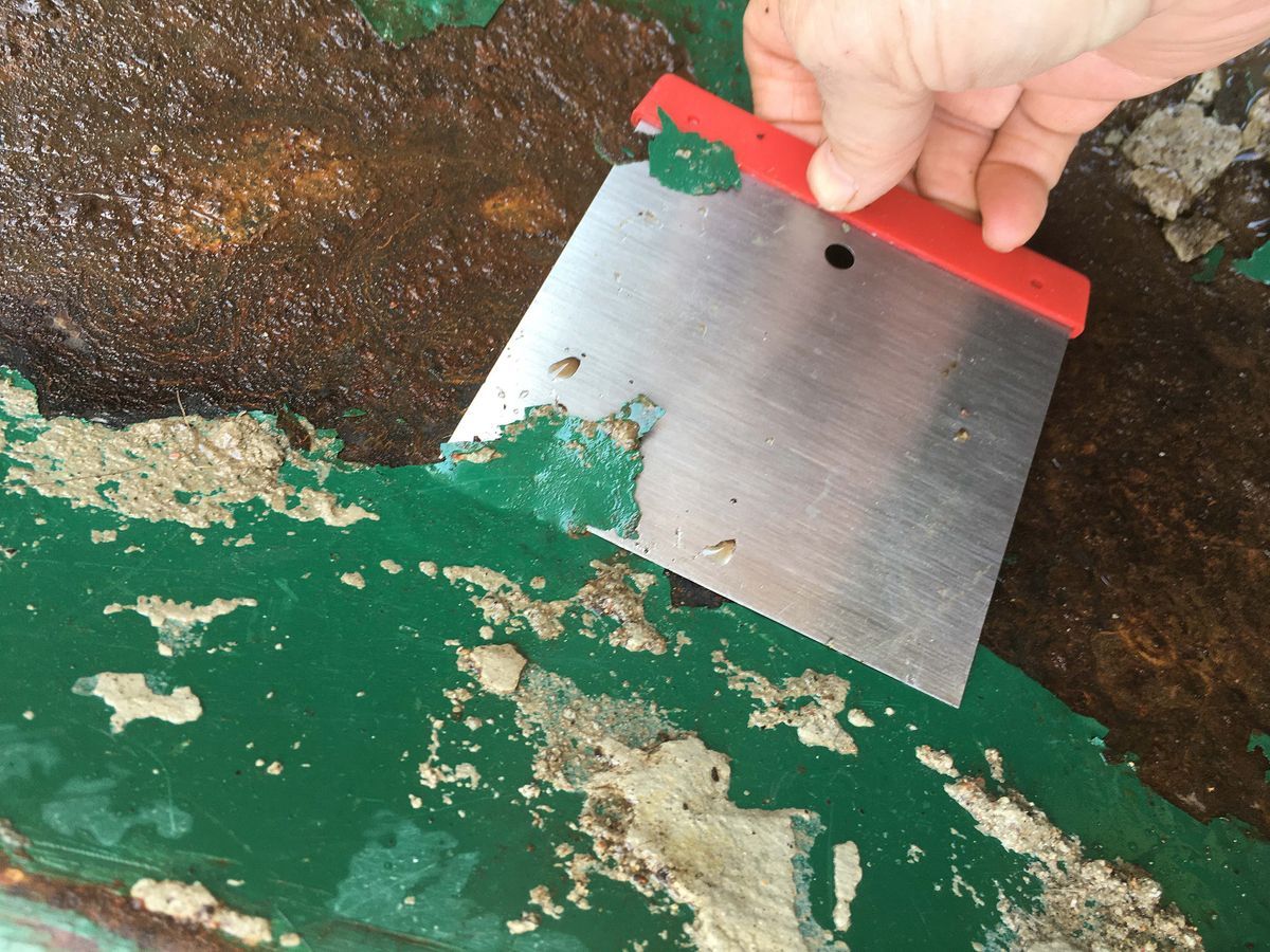 Hand skrapar bort grön färg och rost från en metallisk yta med spackelspade