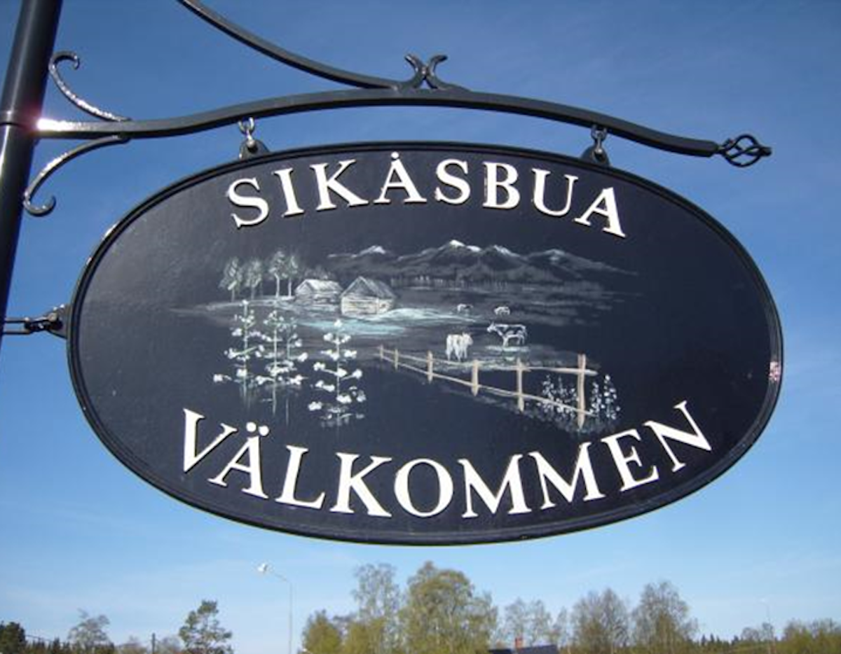 Skylt med texten ”Sikåsbua Välkommen” och landskapsmotiv i bakgrunden